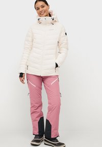 Kvinde iført en hvid hættejakke med dun, lyserøde snebukser med sorte detaljer og sorte sneboots, stående og smilende mod en ensfarvet baggrund.
