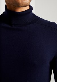 Man som bär en mörk marinblå ribbad turtlenecktröja, som visar detaljer på hals, axlar och övre bröst.