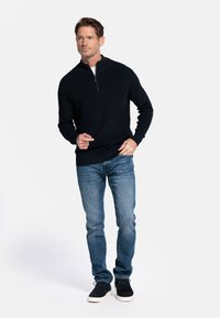 Marineblauwe sweater met ritssluiting in ribbelbreiwerk, gecombineerd met spijkerbroek van gemiddelde wassing en zwarte sneakers. Model staat op een witte achtergrond.