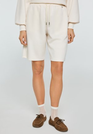 Osoba ubrana w kremowe szorty ze sznurkiem, białe krótkie skarpetki oraz brązowe zamszowe mokasyny na jednolitym tle.