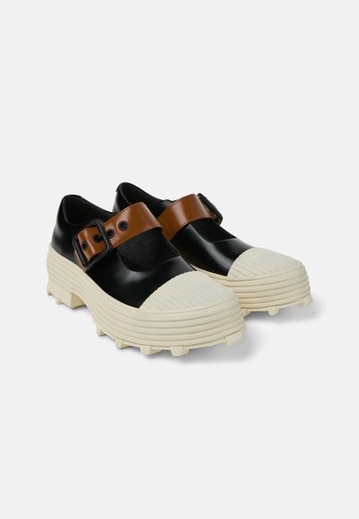 Chaussures noires et brunes avec une semelle en caoutchouc blanc côtelé bien visible, dotées d'une large lanière avec boucle et d'une section découpée sur le dessus.