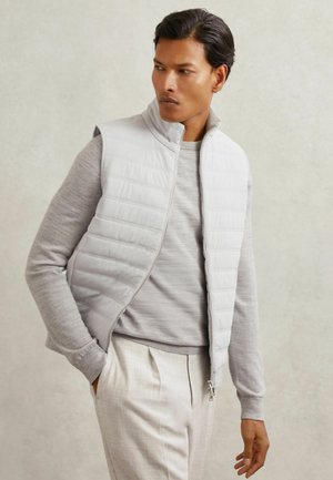Világosszürke puffervest, cipzáros elülső résszel, függőleges steppelt textúrával, magas gallérral, egy szürke, hosszú ujjú póló és világos nadrág fölött viselve.