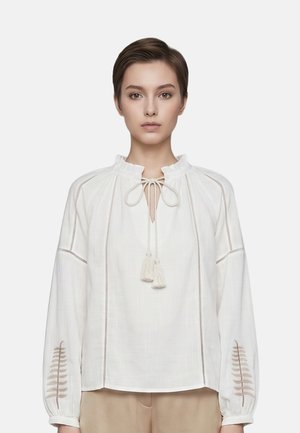 Jonge vrouw met kort haar die een witte blouse met lange mouwen draagt, met tassel-ties en geborduurde varenmotieven op de mouwen.