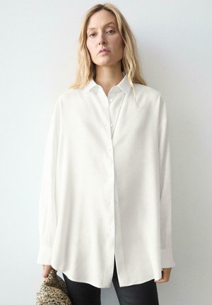 Vrouw met lang blond haar die een oversized wit overhemd en zwarte broek draagt, met een beige clutch met textuur in haar linkerhand.