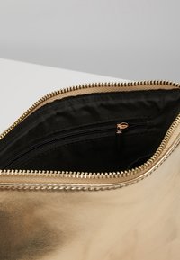 Kultainen metallinhohtoinen pussi, jossa on sileä pinta, vetoketjusulku ja musta kangassisustus lisävetoaukollisella taskulla.