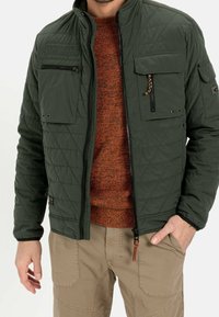 Grøn quiltet jakke med ståkrave, to brystlommer og kontrasterende lynlåse, båret over en orange tekstureret sweater.