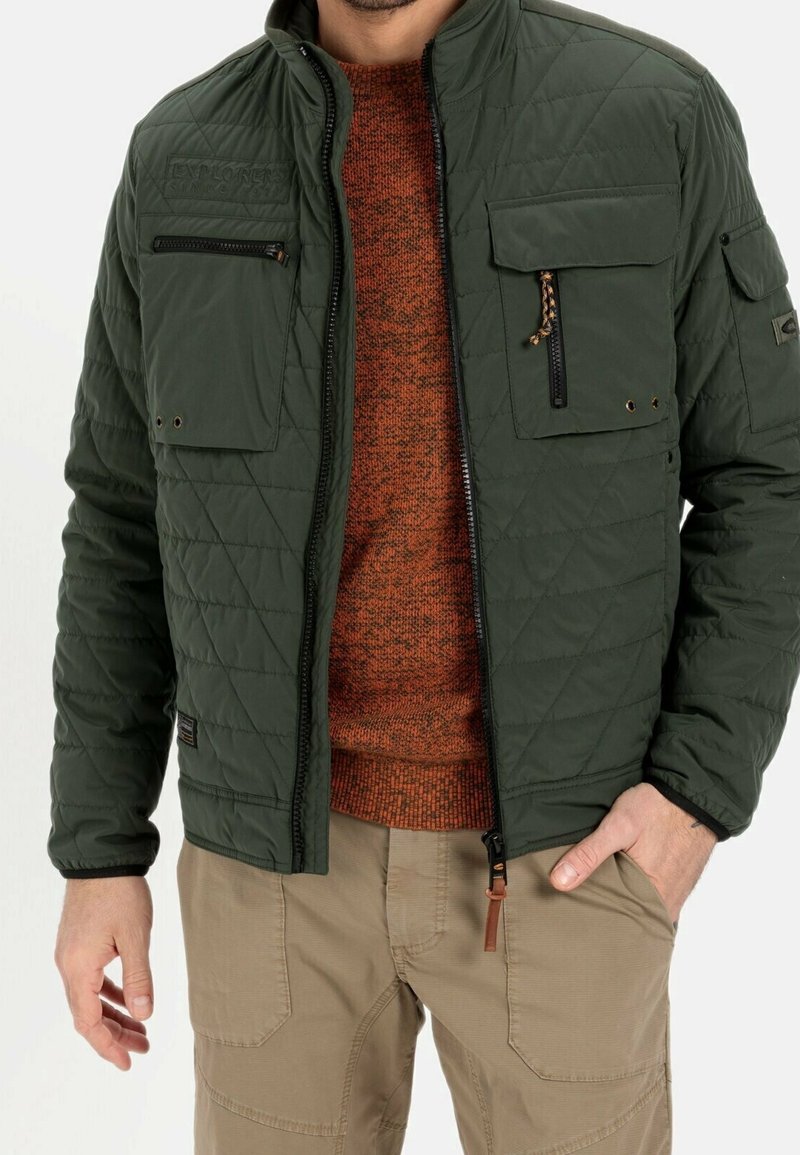 Grøn quiltet jakke med ståkrave, to brystlommer og kontrasterende lynlåse, båret over en orange tekstureret sweater.