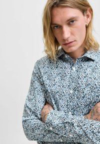 Camicia in cotone con stampa floreale blu. Presenta un colletto aperto, chiusura con bottoni e maniche lunghe con polsini. Motivo floreale dettagliato su tutta la camicia.