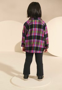 Chemise en flanelle à carreaux multicolores dans les tons de violet, vert et noir, associée à un pantalon extensible noir et des baskets noires, vue de l'arrière.