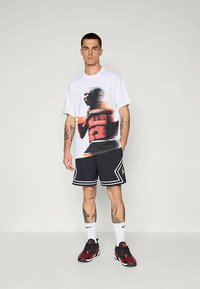 Homem vestido com uma t-shirt de basquetebol gráfica branca, calções de basquetebol pretos com contorno branco, meias Nike brancas e ténis vermelhos e pretos, de pé.