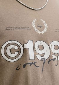 Brązowy T-shirt bawełniany z wyraźnym nadrukowanym tekstem i dekoracyjnym wzorem laurowym. Wyraźne przetarcia na tekście i fakturze materiału.