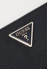 Must värviline rahakott, millel on must tekstuur ja kolmnurkne metalne logo kuldses värvitoonis koos tekstiga "GUESS USA" ning küsimärkide kujundusega.