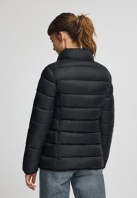 Svart pufferjacka sedd bakifrån, med hög krage, horisontell sömnad och en slät, glansig yta. Figursydd design.