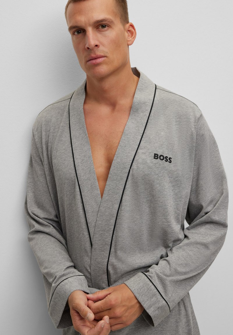 Robe grise avec un passepoil noir, faite de tissu doux. Elle possède un col châle et des manches longues, avec une ouverture sur le devant et un logo brodé sur la poitrine.