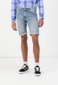 Valkoiset denim-shortsit vaaleansinisellä sävyllä, jossa on revitty helma, käännetyt hihnat ja klassinen viisi taskua -tyyli, yhdistettynä mustiin kenkiin ja valkoisiin sukkiin.