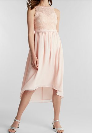 Kobieta w bezrękawną, jasnoróżową sukienkę o asymetrycznym kroju z koronkowym gorsetem, stojąca z rozstawionymi nogami, w srebrnych szpilkach z paskiem wokół kostki na białym tle.