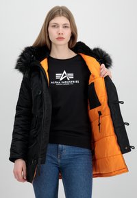 Crna puf jakna s kapuljačom i umjetnim krznom. Unutra je svijetlo narančasta, s patentnim zatvaračima i dugmadima. Model nosi crnu duksericu.