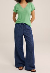 Žena oblečená v světle zeleném topu s výstřihem do V, s volnými tmavě modrými kalhotami na šnůrku a hnědými sandály, stojící s rukama za zády.