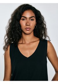 Vrouw met krullend zwart haar, gekleed in een zwart mouwloos topje en grote ronde oorbellen, kijkt recht vooruit tegen een effen lichte achtergrond.