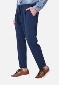 Pantaloni jogger navy con vita elasticizzata, lacci neri, tasche laterali e gambe affusolate, abbinati a scarpe da ginnastica marroni con lacci.