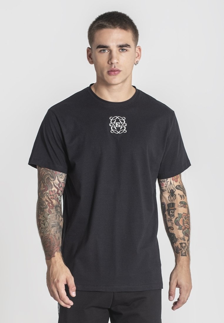 Svart bomullströja med ett framåtriktat grafiskt tryck i vitt, rund hals, korta ärmar och en avslappnad passform. Tydliga arm-tatueringar synliga.
