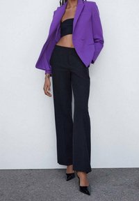Blazer violet aux épaules structurées, crop top noir, pantalon noir évasé taille haute, et escarpins noirs à bout pointu.
