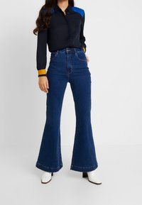 Camiseta navy con hombros en bloques de color azul y naranja, combinada con pantalones vaqueros de tiro alto, acampanados y de color azul oscuro, y botas de tacón blancas.