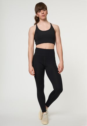 Noor naine seisab ühe jalaga kõverdatuna, seljas must spordirinnahoidja, mustad kõrge vöökohaga retuusid ja beežid tossud ühevärvilisel taustal.