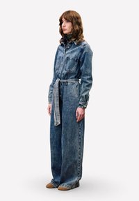 Denim jumpsuit met een knoopsluiting aan de voorkant, twee borstzakken, ruime benen en een gestrikte riem. Draagt met bruine instappers.
