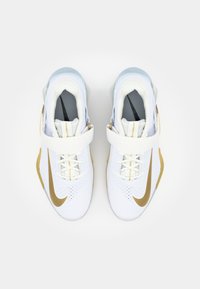 Zapatos deportivos blancos con un swoosh de Nike dorado, parte superior texturizada, cuello acolchado y una correa en el empeine para mayor soporte.