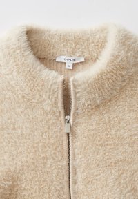 Beige, flauschiger, strukturierter Reißverschluss-Pullover mit silbernem Reißverschluss und einem kleinen weißen Etikett im Kragen mit der Aufschrift "OPUS 36."