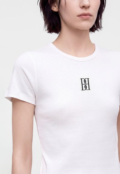 Femme portant un t-shirt blanc uni à manches courtes avec un petit logo monogramme noir centré sur la poitrine.