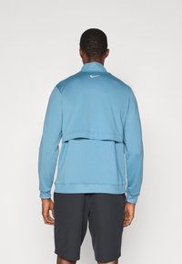 Sweatshirt Nike de culoare albastră, cu guler, mâneci lungi și un design stratificat. Prezintă un mic logo alb pe spate și o textură moale.