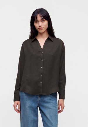 Femme aux longs cheveux foncés portant une chemise marron foncé boutonnée et un jean bleu sur un fond clair uni.