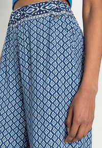 Modrý a bílý vzorovaný látkový materiál s geometrickým designem. Elastický pas, uvolněný střih, měkká textura, zobrazený na ruce vedle oděvu.