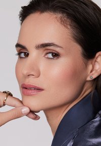 Zbliżenie na osobę z gładką skórą, noszącą złoty pierścień i małą niebieską kolczyk, ukazaną w neutralnym makijażu i ciemnych włosach w satynowej kurtce.