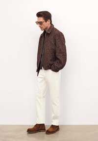 Giacca bomber marrone con texture sopra una camicia a bottoni scura, abbinata a pantaloni bianchi e stivali stringati marroni. Gli occhiali da sole completano il look.