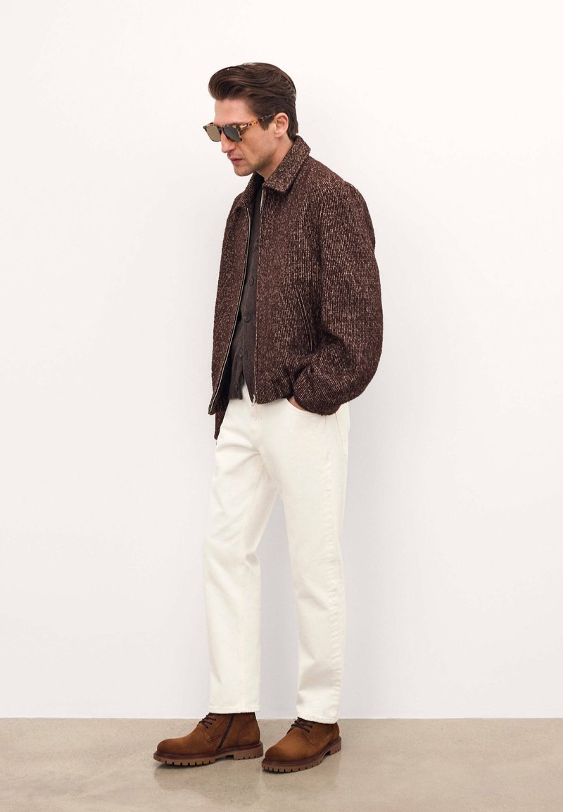Jachetă bomber texturată maro peste o cămașă închisă cu nasturi, asortată cu pantaloni albi și cizme maro cu șireturi. Ochelarii de soare completează look-ul.