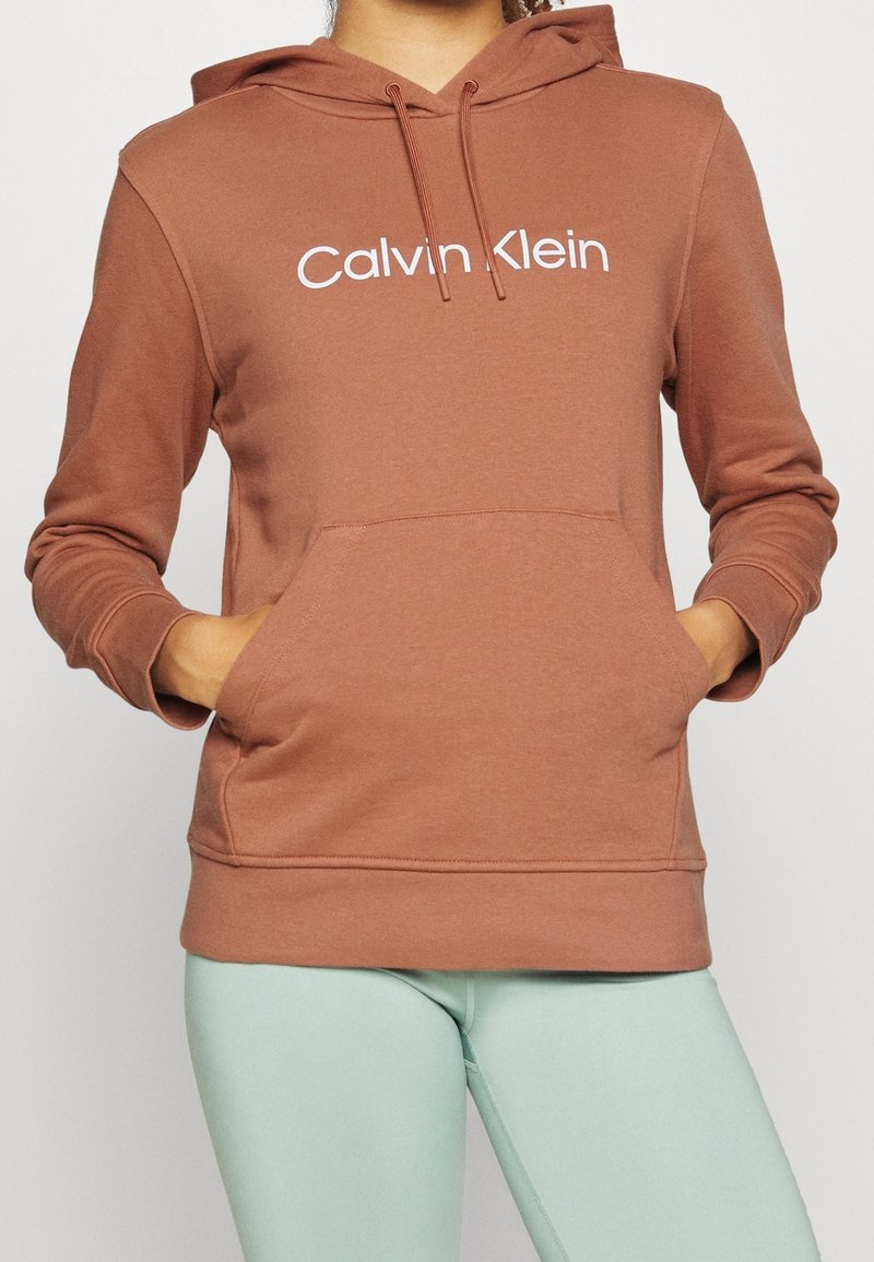 Brun hættejakke lavet af blødt stof med en frontlomme, prydet med et hvidt "Calvin Klein" logo på brystet og en snøre i hætten.