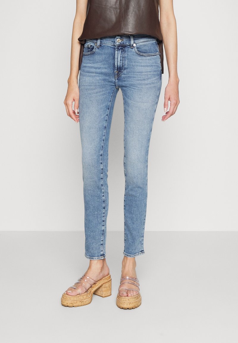 Femme portant un jean slim bleu clair et des sandales à plateforme en liège avec brides transparentes, debout devant un fond clair uni.