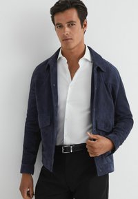 Chaqueta de ante azul marino con cuello, bolsillos frontales y botones a presión, llevada sobre una camisa blanca y pantalones negros. Textura suave.