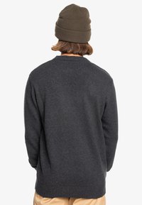 Mörkgrå stickad tröja med en strukturerad yta och ribbad nederkant. Bärs med en brun mössa, som visar en enkel, ogrillad design.