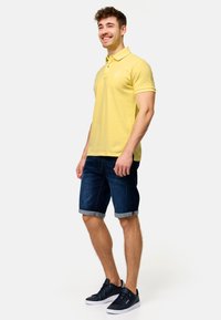 Gul polo t-shirt med krave, korte ærmer, sammen med mørk denimshorts, der er rullede ved kanten, og mørke sneakers.