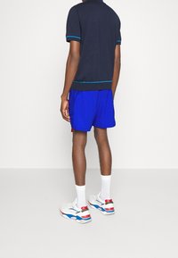 Maglietta polo blu navy con rifiniture blu, abbinata a pantaloni corti blu. Sneakers bianche con accenti rossi, blu e dorati. Sfondo semplice.