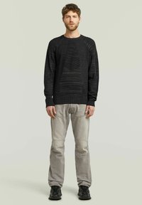 Pull en tricot noir texturé avec des manches raglan, associé à un jean gris clair et des baskets noires, se tenant sur un fond neutre.