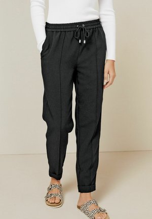 Frau trägt schwarze Hose mit Kordelzug und Seitentaschen, weißes geripptes Langarmshirt und Sandalen mit gepunkteter Schnalle, steht auf einfachem Boden.