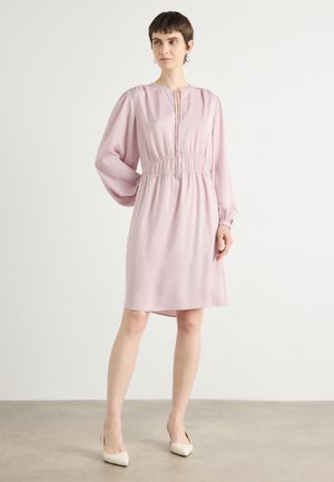 Robe rose clair en tissu lisse, dotée de longues manches bouffantes, d'une taille froncée et d'un décolleté en trou de serrure. Portée avec des talons blancs.