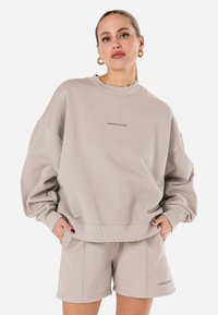 Beige oversized sweatshirt med ribbestrikket hals og senkede skuldre, kombinert med matchende shorts med sidelommer og logo-detalj.