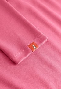 Opgevouwen mouw van een roze kledingstuk met een klein oranje label met een witte honden-silhouet vlakbij de boord.