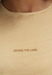 Beige T-shirt with a round neckline and short sleeves. Features "SENSES. THE LABEL" embroidered in a darker beige. Soft, smooth texture.